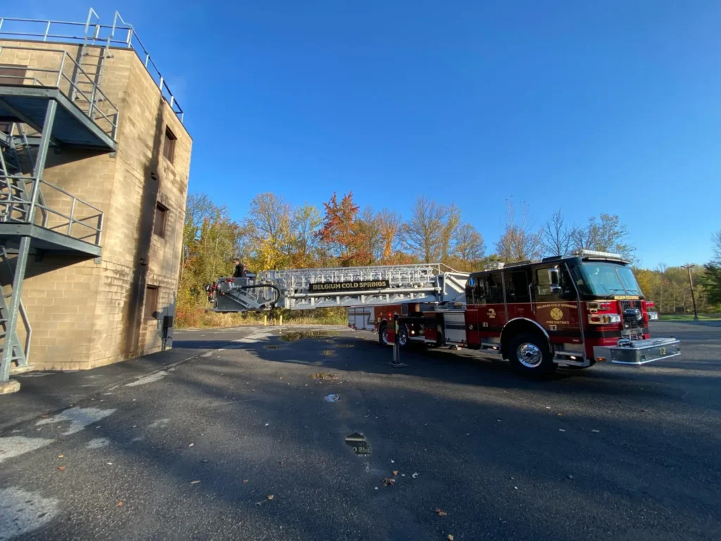 A Belgium Cold Spring fire truck parked next to a tall building using it's extendable ladder